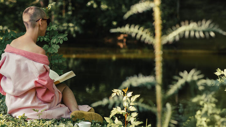 A woman with short hair and bald spots sitting by the lake and reading a book